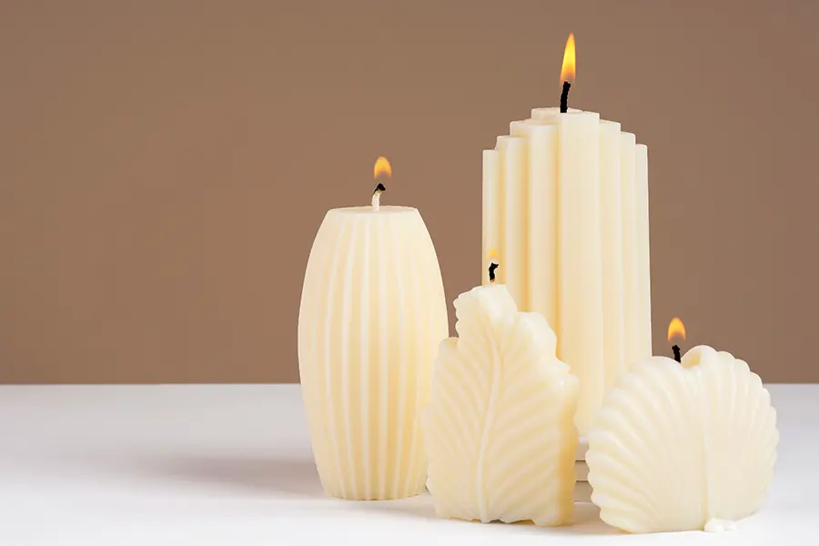 Candele decorative cilindriche e a colonna disposte su una mensola in legno chiaro accanto a elementi naturali come rami secchi e ceramiche artigianali